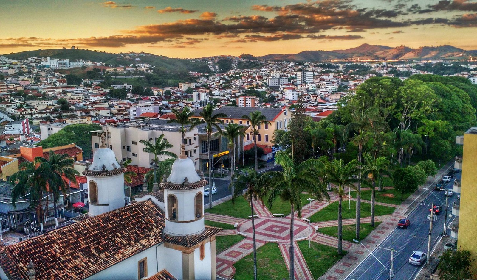 Vista aérea do centro de Campo Belo ao entardecer, com as torres da igreja matriz e a praça arborizada